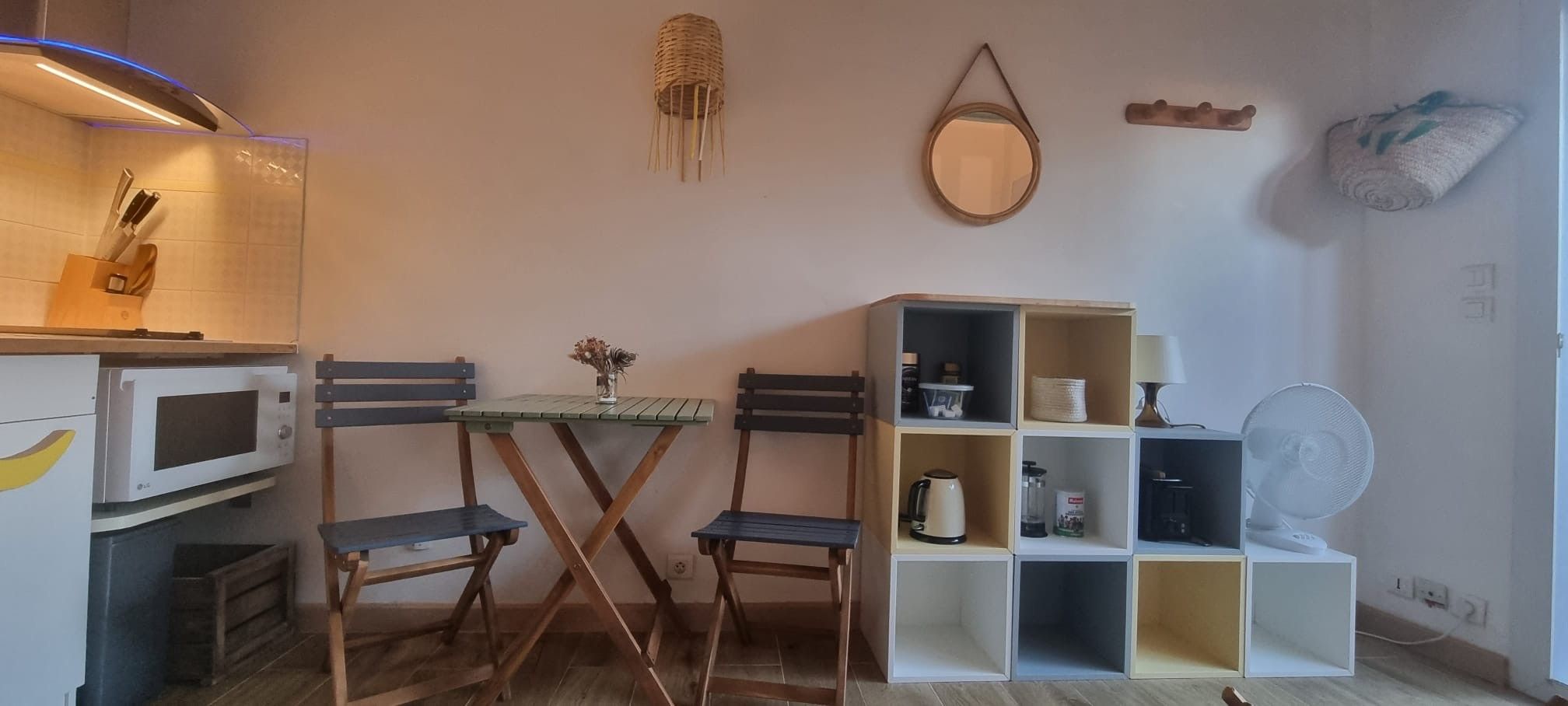 A compact kitchen with a microwave, a small dining table with chairs, and a shelf unit with various items.