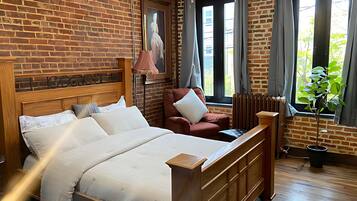 Habitación de hotel con pared de ladrillo, cama con sábanas blancas, un sillón rojo, un radiador y una ventana con cortinas.