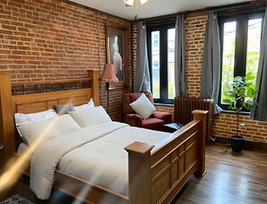 Quarto de hotel com parede de tijolos, cama com roupa de cama branca, poltrona vermelha, radiador e janela com cortinas.