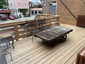 Um deck de madeira com mesa e cadeiras, um muro de tijolos e vista para uma rua residencial.