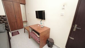 Una habitación compacta con un escritorio de madera, un televisor fijado a la pared, un armario y un pequeño cubo de basura.