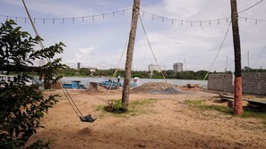 Zona arenosa de playa con un columpio, un bote amarrado junto al agua y materiales de construcción al fondo.