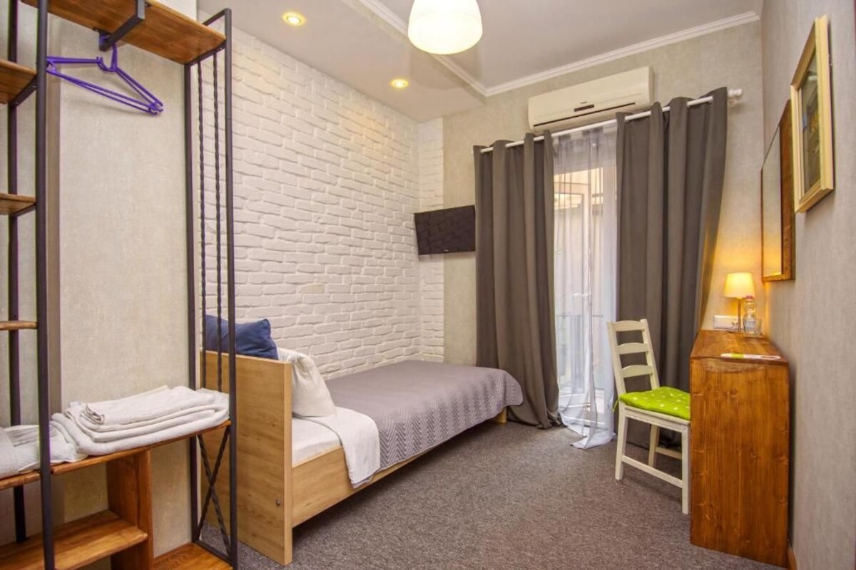 Een kleine slaapkamer met een bed, een stoel, een nachtkastje, een kledingrek en een televisie aan de muur.