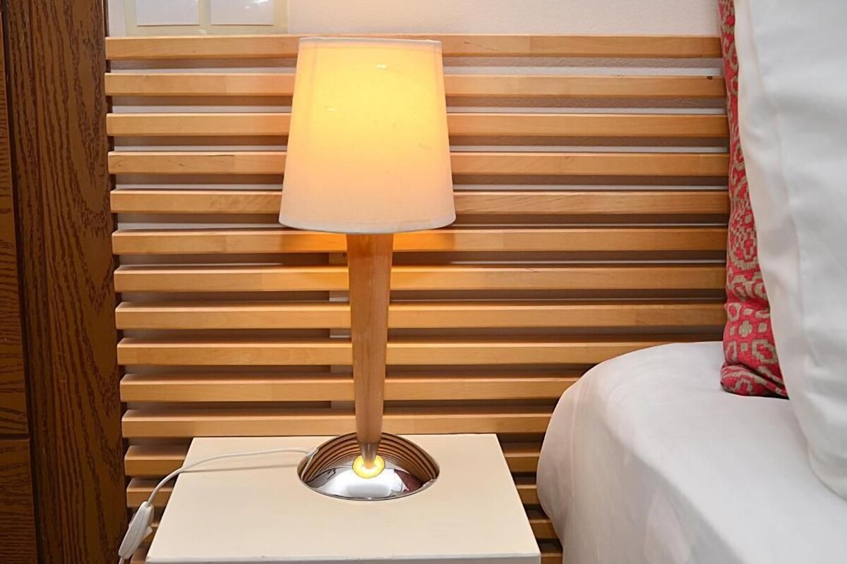 Een nachtkastje met een lamp, een bed met wit beddengoed en een houten lattenbovenbord.