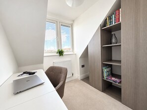 Une petite pièce bien éclairée, avec un bureau blanc, un ordinateur portable, une chaise marron, un radiateur et une bibliothèque contenant des livres et des objets décoratifs.