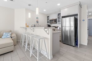 Une cuisine moderne équipée d’un réfrigérateur en acier inoxydable, d’un four à micro-ondes et d’une machine à café. On y trouve trois tabourets de bar blancs et un canapé blanc agrémenté d’un coussin bleu.