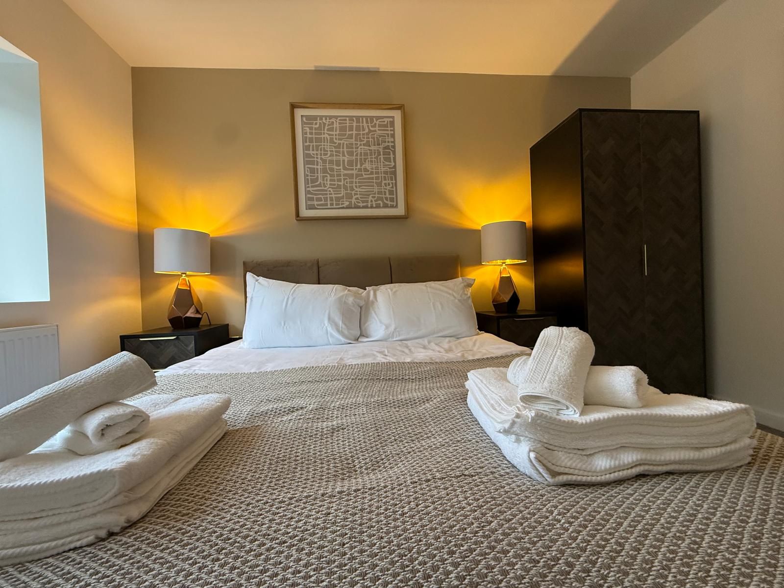 A hotel room with a bed, two bedside lamps, a large dark wardrobe, and a folded towel on the floor.