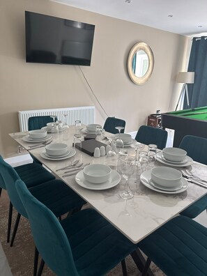 Uma sala de jantar com uma mesa posta para quatro, cadeiras, uma televisão na parede e uma mesa de bilhar ao fundo.