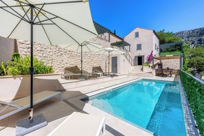 Une piscine avec des chaises longues et des parasols, un mur en pierre et un bâtiment doté d’un escalier.