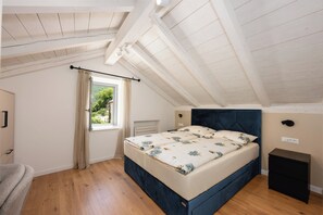 Une chambre avec un plafond en pente, un lit recouvert d’une literie à motifs floraux, une fenêtre avec des rideaux et une petite table de chevet.