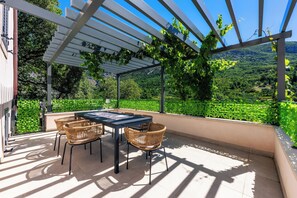 Un espace extérieur abrité, doté d’une table et de chaises, entouré de verdure et offrant une vue sur un paysage vallonné.