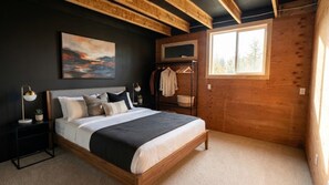 Une chambre à coucher comprenant un lit, des tables de chevet, une commode, un téléviseur fixé au mur, un porte-manteau et une fenêtre donnant sur la forêt.