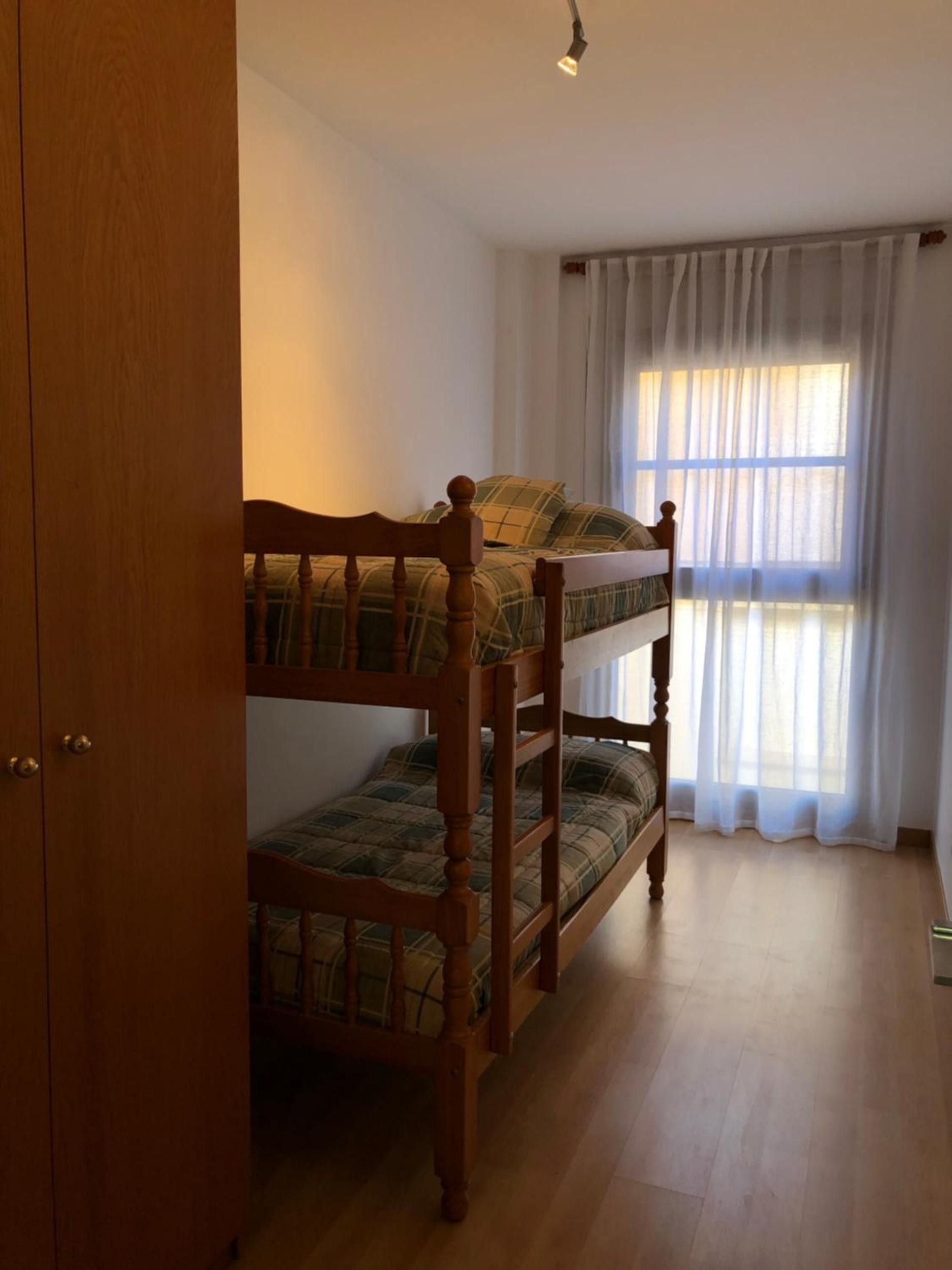 Un lit superposé avec une structure en bois et une literie à carreaux, une armoire en bois et une fenêtre ornée de rideaux légers.