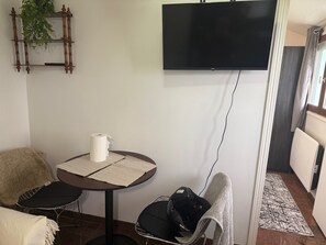 Una habitación con una televisión de pantalla plana montada en la pared, una mesa redonda con un rollo de papel higiénico, una silla y un abrigo colgado sobre otra silla.