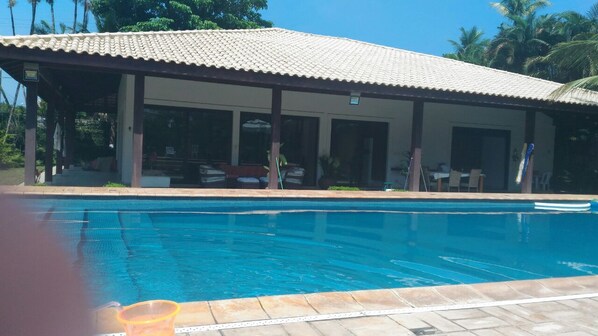 Piscina com área coberta, palmeiras e céu azul.