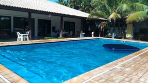 Piscina com deck ladrilhado, área de estar coberta e palmeiras ao fundo.