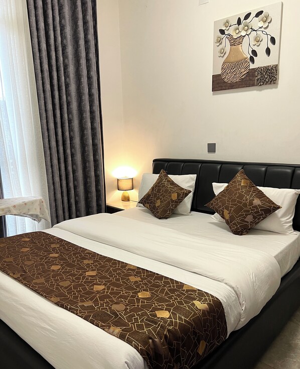 A hotel room with a bed, a nightstand with a lamp, a painting on the wall, and a window with curtains. - CosyNook apartments (Kigali)
