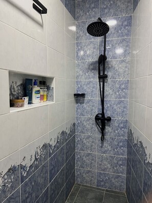 A shower with a handheld showerhead, a shelf with toiletries, and a tiled wall with a floral pattern. - CosyNook apartments (Kigali)