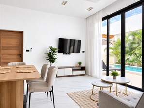 Un salon moderne avec une table à manger en bois, deux chaises, un canapé, un téléviseur et une vue sur une piscine.