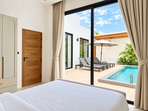Une chambre d’hôtel avec un grand lit et une porte-fenêtre coulissante donnant sur un espace piscine équipé de chaises longues et de parasols.