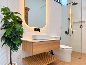 Une salle de bain moderne avec un lavabo blanc, un meuble-lavabo en bois, un miroir, une douche et une plante en pot.