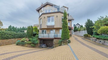 A multi-story residential building with a balcony, a stone wall, and a paved driveway.