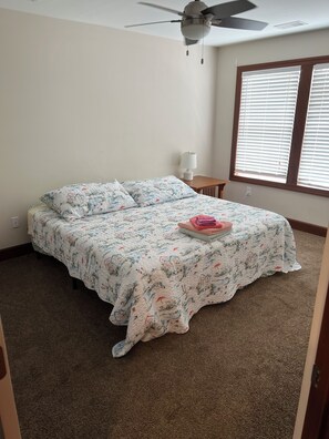 Un dormitorio con cama, ventilador de techo, ventana con persianas y lámpara en una mesita de noche.