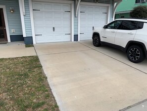 Un garaje con dos puertas blancas, una camioneta SUV blanca estacionada adentro y un camino de hormigón que conduce al garaje.