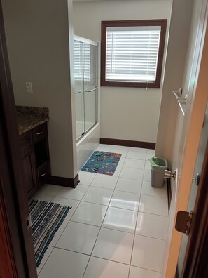 Un baño con ducha, una ventana con persianas, piso de baldosas y un cubo de basura.