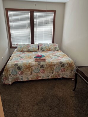 Une chambre à coucher avec un lit, deux fenêtres équipées de stores et un sol recouvert de moquette.