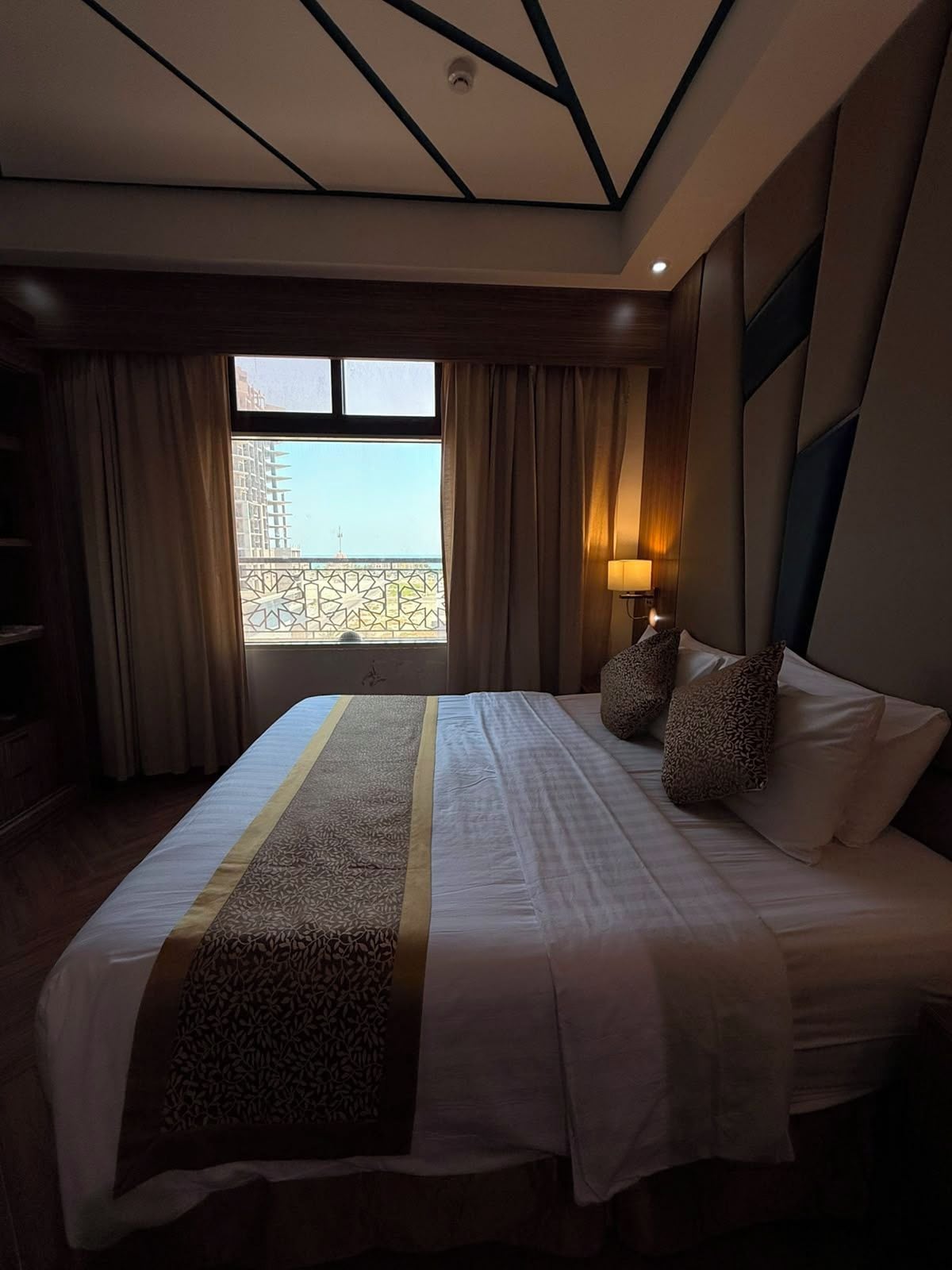 A hotel room with a large bed, a view of a building through a window, and a nightstand with a lamp.