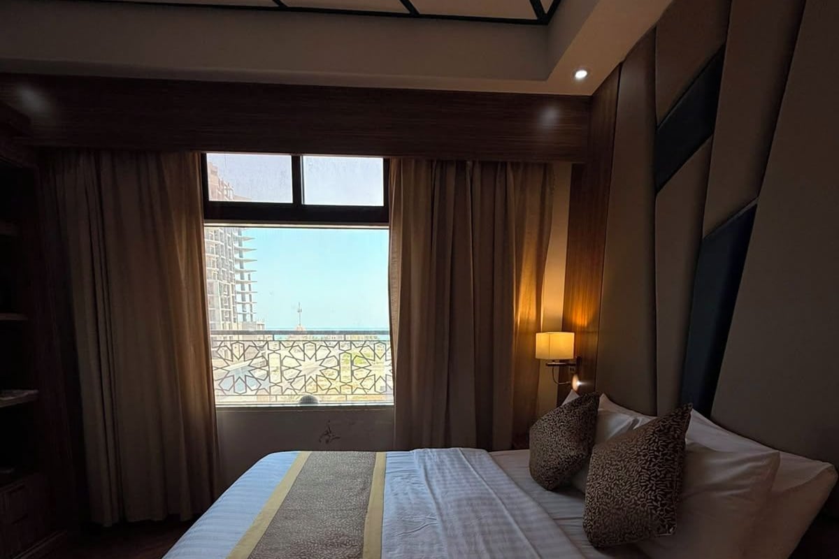 A hotel room with a large bed, a view of a building through a window, and a nightstand with a lamp.