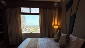 A hotel room with a large bed, a view of a building through a window, and a nightstand with a lamp.