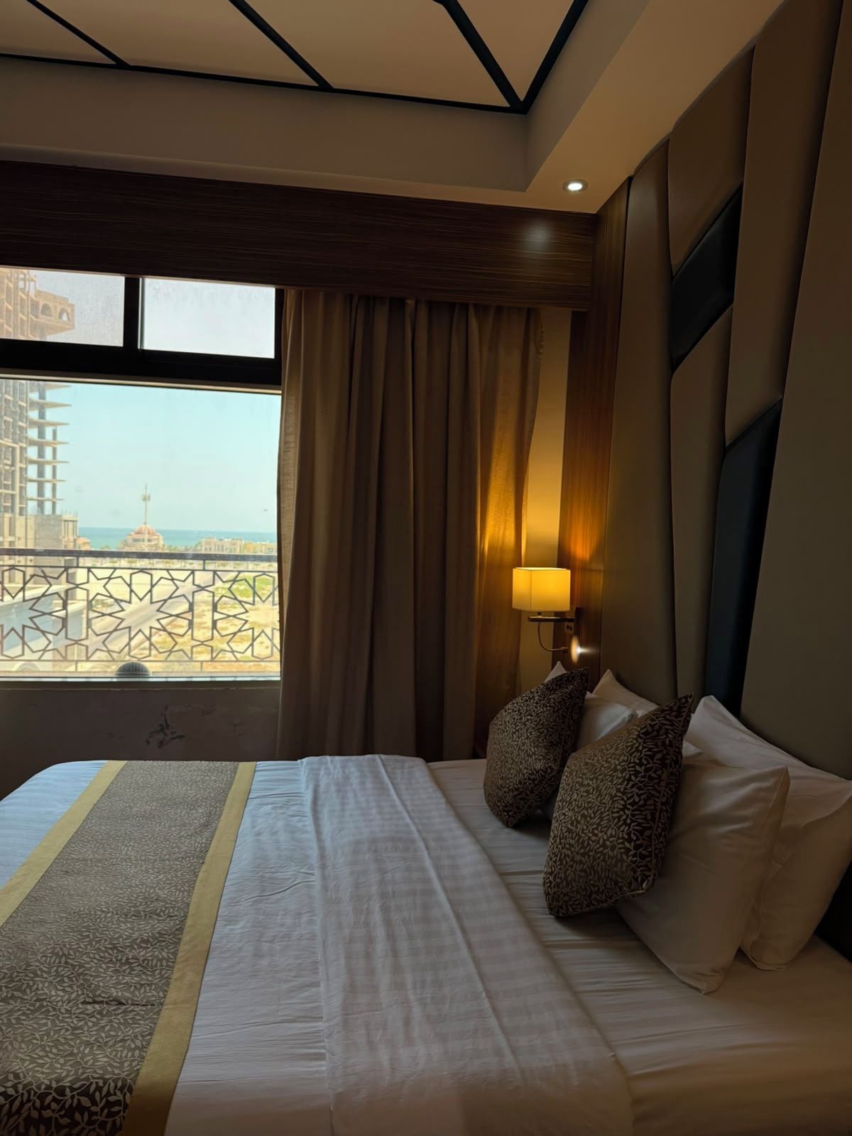 A hotel room with a bed, a window with curtains, a view of a building, and a lamp.