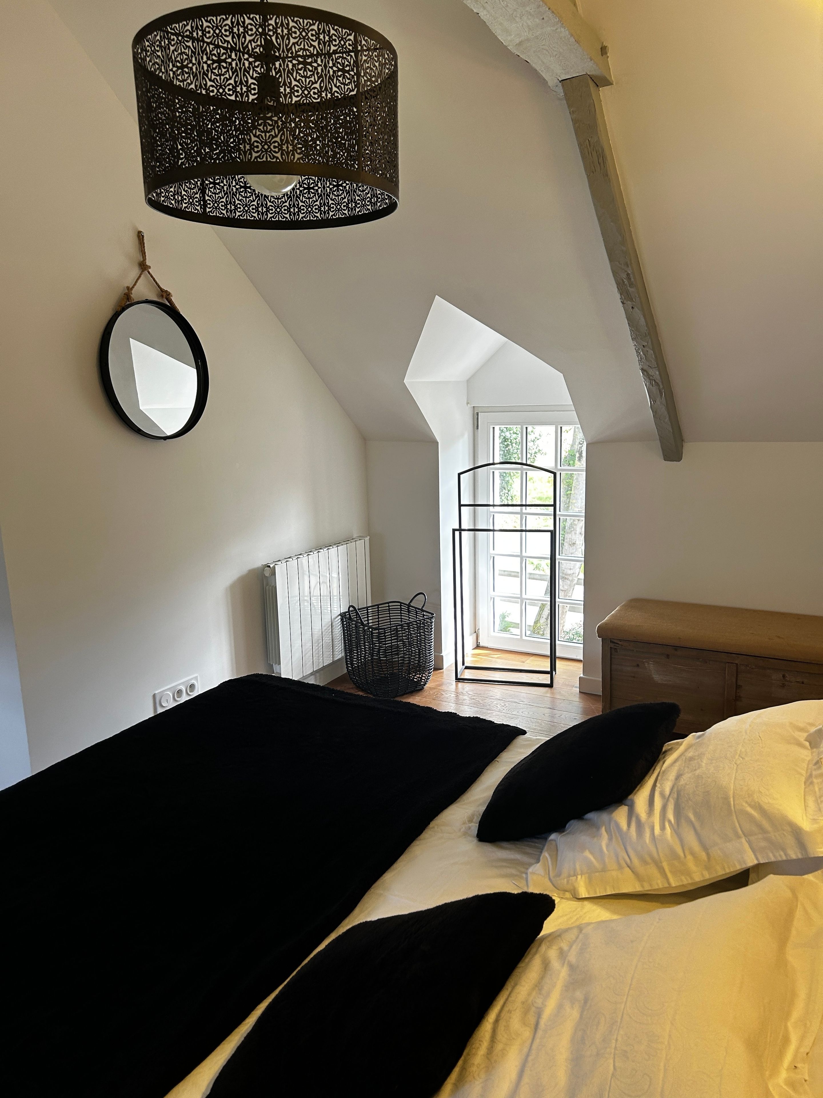 Una camera da letto con soffitto spiovente, un letto con biancheria a pois bianchi e neri, una lampada a sospensione con motivo a scacchi bianco e nero, uno specchio tondo e una finestra con vista su piante.