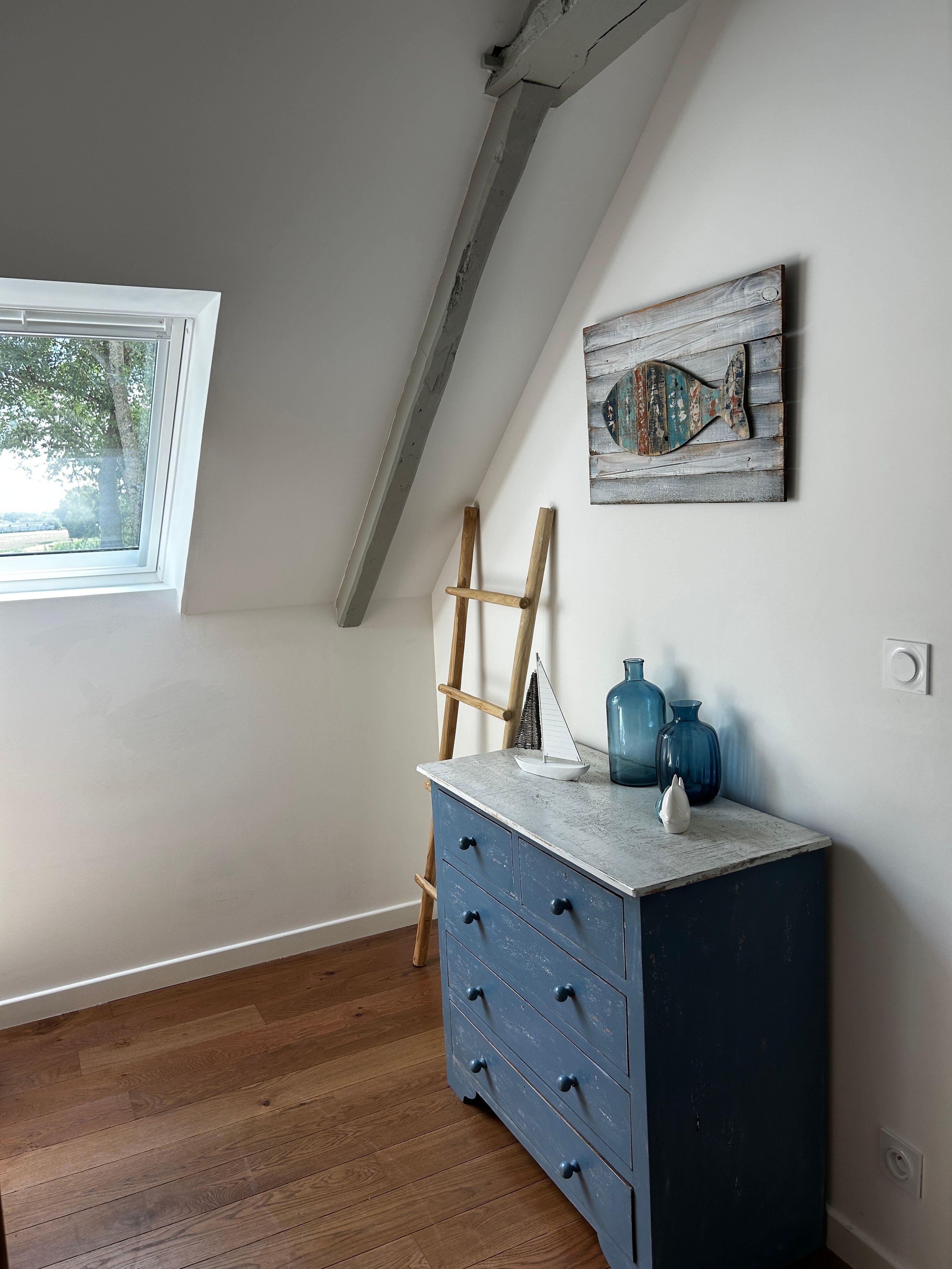 A room with a sloped ceiling, a window, a dresser with drawers, a ladder, and a wall-mounted artwork.