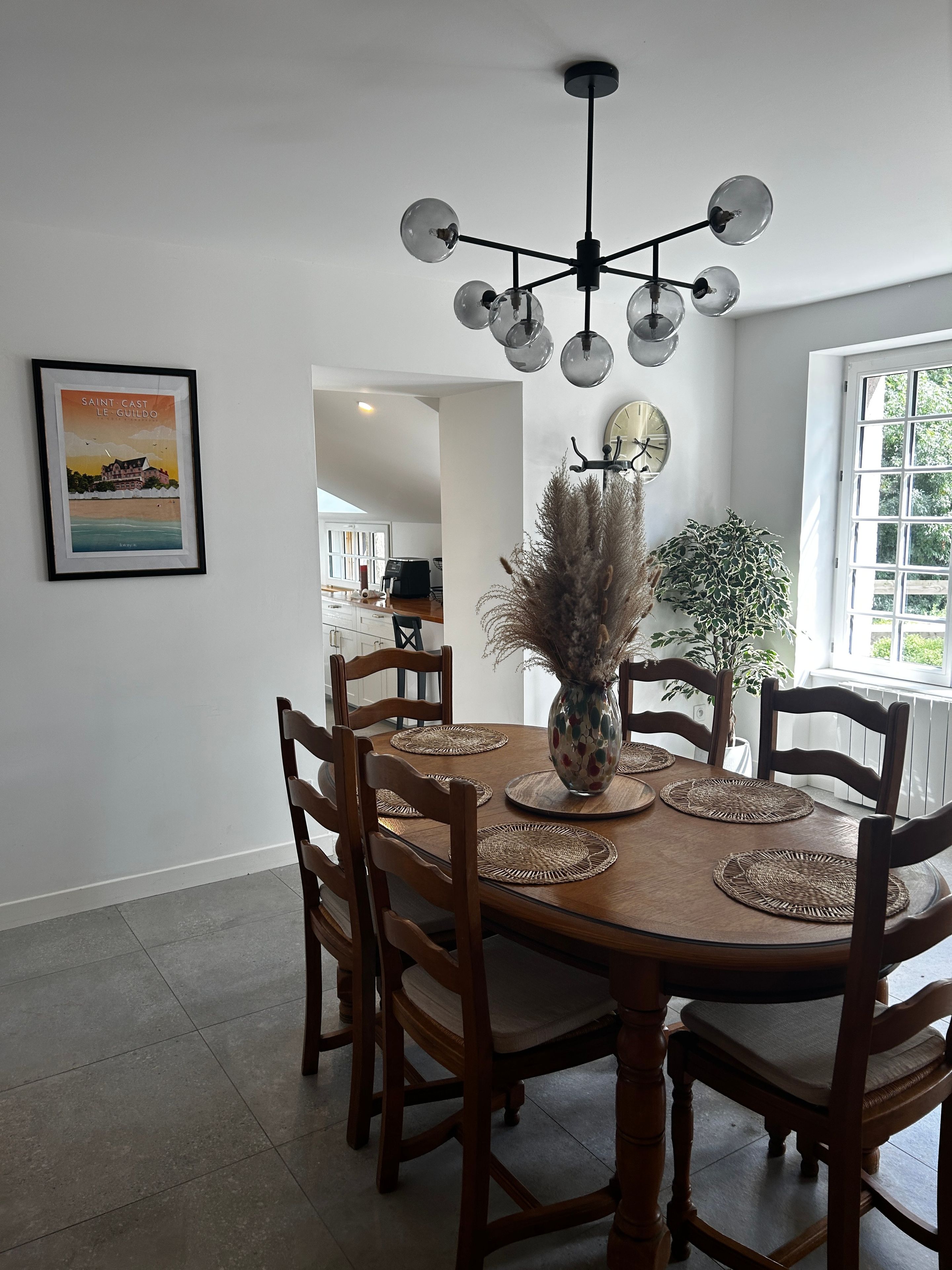 A dining area with a round table, wooden chairs, a vase with dried plants, a wall-mounted clock, and a framed picture.