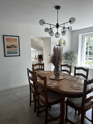 Een eetruimte met een ronde tafel, houten stoelen, een vaas met gedroogde planten, een klok aan de muur en een lijst met een afbeelding.