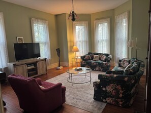 En stue med et fladskærms-tv, en rød lænestol, en blomstret sofa og en lampe.