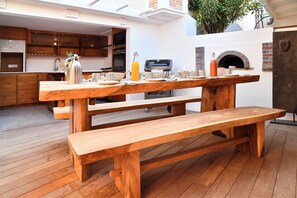Ein hölzerner Essbereich im Freien mit Bank und Tisch, einem Grill und einem Backofen aus Ziegeln.