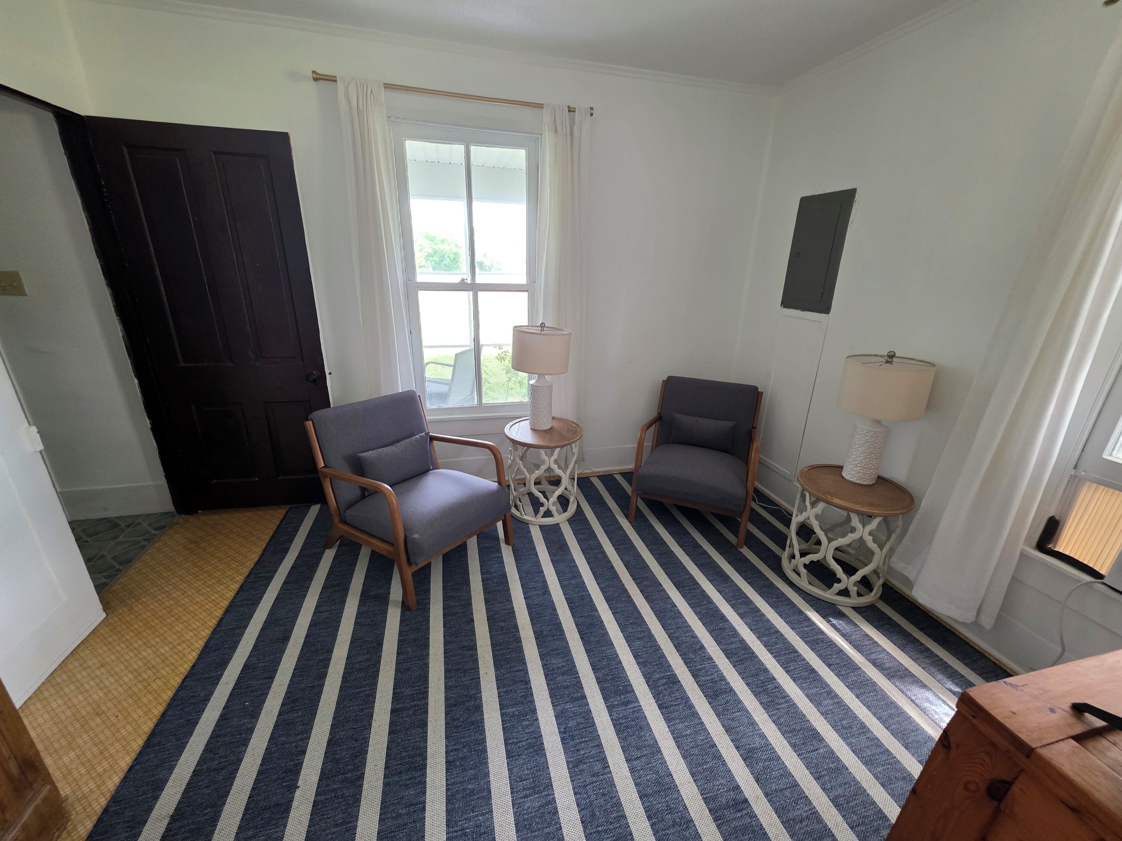 A room with a striped carpet, two chairs, a small table with a lamp, and a window with curtains.