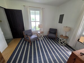 Une pièce avec un tapis à rayures, deux chaises, une petite table avec une lampe et une fenêtre avec des rideaux.