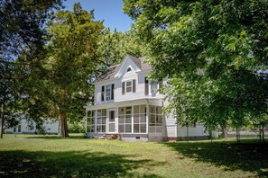 Une maison à deux étages avec un porche, entourée d’arbres et dotée d’une cour clôturée.
