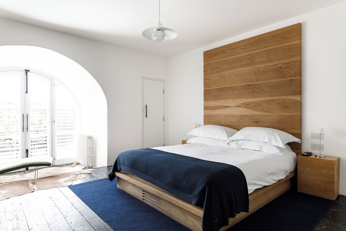 A modern bedroom with a wooden headboard, a bed with white linens, a blue rug, and a nightstand with a lamp.