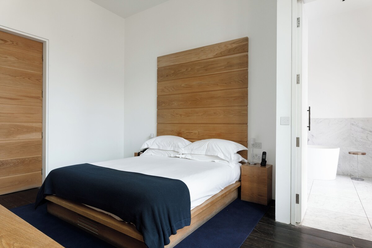A modern bedroom with a wooden headboard, a single bed with white bedding, a bedside table with a lamp, and a bathroom visible through an open door.