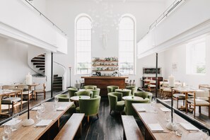 A restaurant with a central bar, green upholstered chairs, wooden tables, and a spiral staircase. - At The Chapel (Bruton)