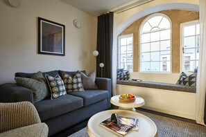A modern living room with a sofa, a side table with a bowl of fruit, and a window with a view of buildings. - At The Chapel (Bruton)