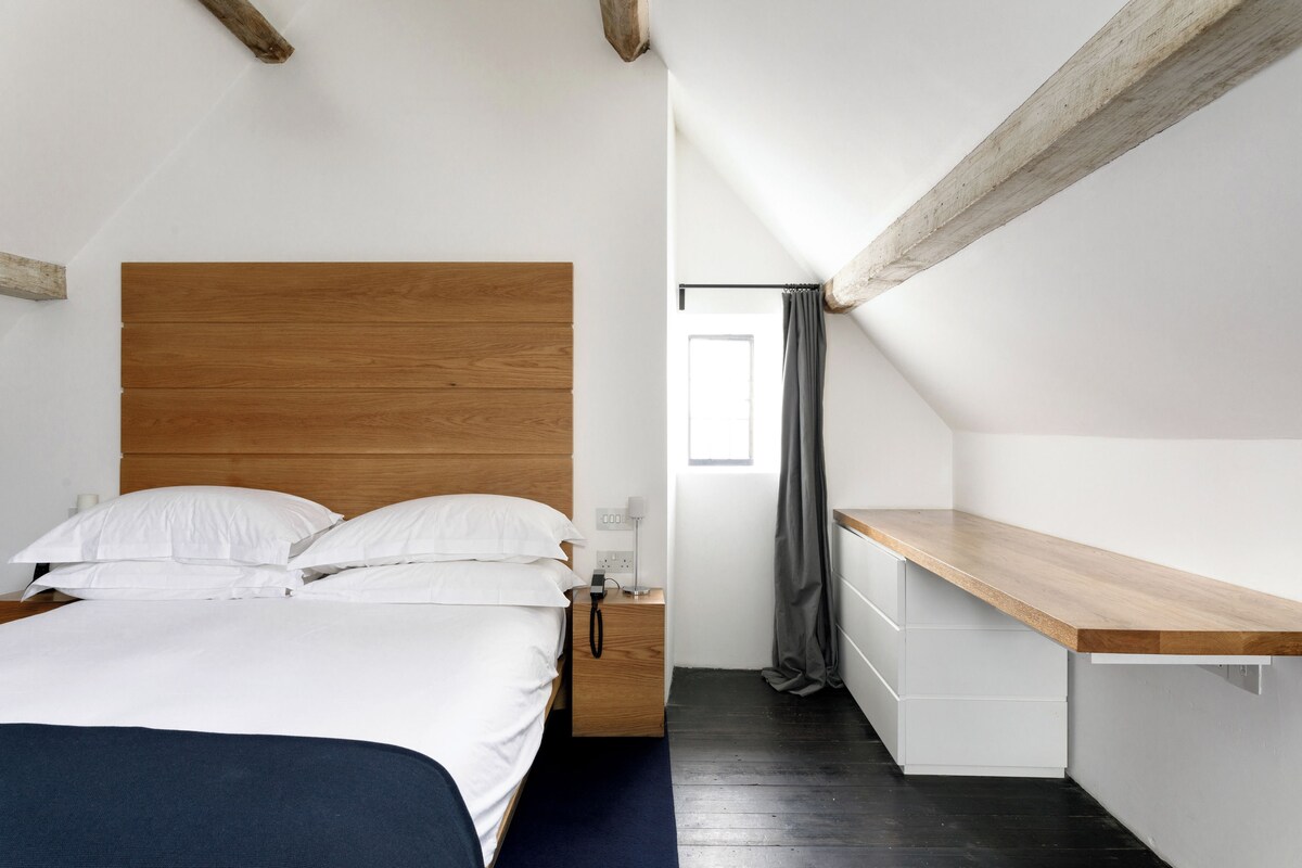 A bedroom with a wooden headboard, a bed with white linens, a bedside table with a lamp, and a window with a curtain.