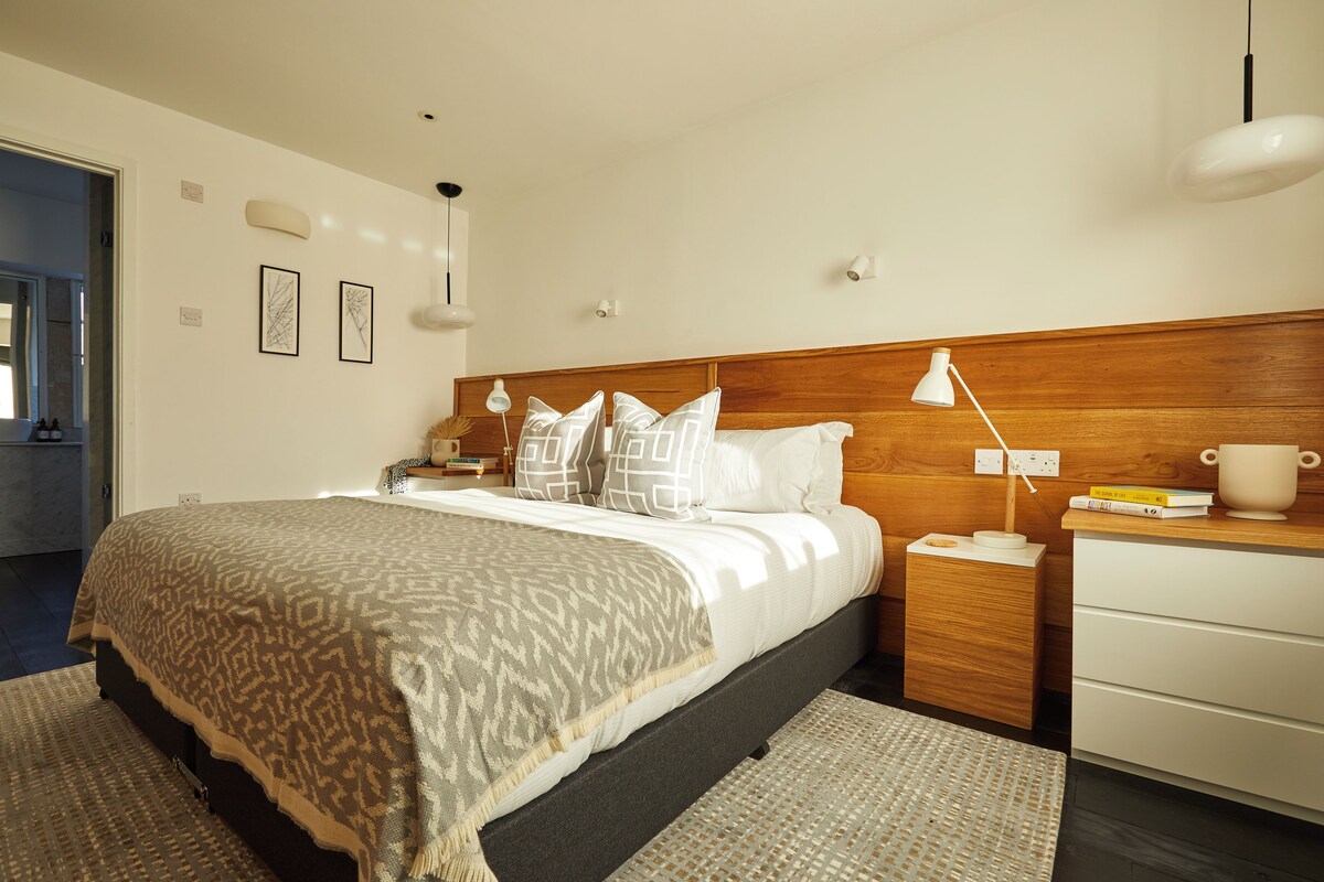 A modern bedroom with a wooden headboard, a bed with patterned bedding, bedside lamps, a nightstand, and a small table with books and a cup.