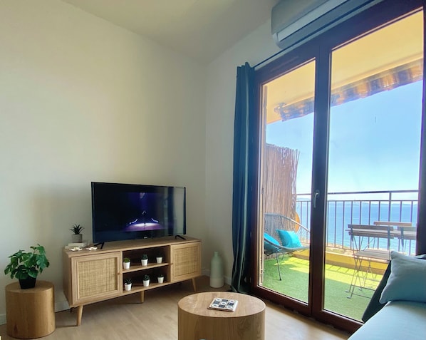 Ein Wohnzimmer mit Flachbildfernseher, Holzschrank, rundem Couchtisch und Balkon mit Gartenmöbeln.